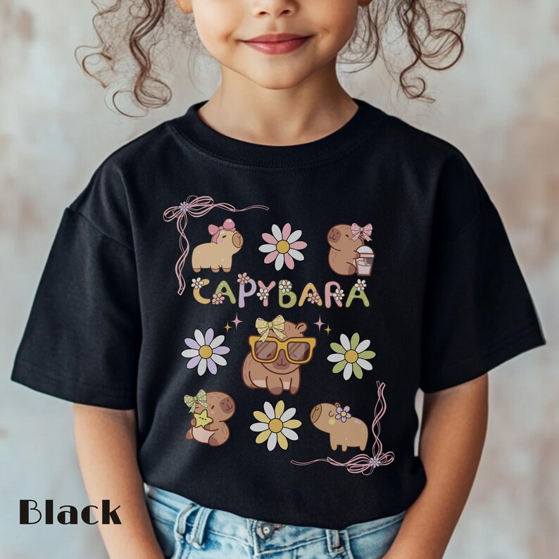 May include: Black t-shirt featuring a colorful graphic design with the word "CAPYBARA" and illustrations of capybaras, flowers, and bows. The shirt is made of a soft material and is suitable for children.