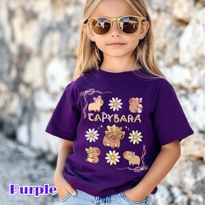 May include: A purple t-shirt featuring a graphic of capybaras with sunglasses, bows, and flowers. The word "CAPYBARA" is printed in a playful font. The shirt is made of a soft material.
