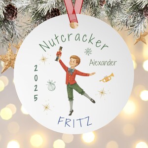 May include: A white Christmas ornament with a red ribbon features a watercolor illustration of a boy holding a nutcracker. The ornament has the text "Nutcracker", "Alexander", "Fritz", and the year "2025".