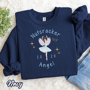 May include: Navy blue sweatshirt with a ballerina angel design. The design includes the text "Nutcracker 2025 Angel", a ballerina in a light blue tutu, angel wings, and gold stars. The word "Navy" is written in cursive at the bottom.