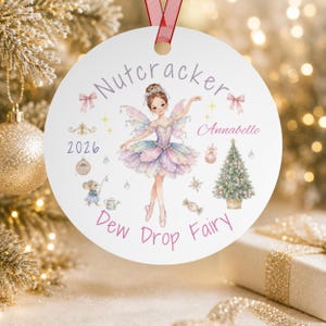 May include: A round white ceramic ornament featuring a fairy in a pastel tutu, a Christmas tree, and a small mouse. The text reads Nutcracker, Annabelle, 2026, and Dew Drop Fairy. It includes pink bows, stars, and a red ribbon for hanging.