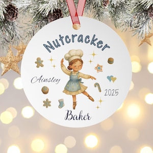 May include: A white Christmas ornament with a red ribbon features a ballerina baker illustration. The ornament has the words "Nutcracker," "Ainsley," and "Baker" in blue script. Decorative gingerbread men, stars, and candy canes surround the ballerina.