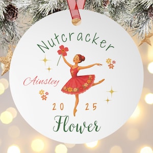 Puede incluir: Adorno navideño redondo blanco con una cinta roja. El adorno presenta una bailarina con un vestido rojo sosteniendo una flor, con el texto "Nutcracker", "Ainsley", "20 25" y "Flower" en varias fuentes y colores.