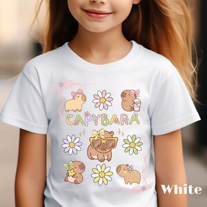 May include: White t-shirt featuring a colorful graphic design with the word "CAPYBARA" and illustrations of capybaras, flowers, and bows. The design includes various colors such as yellow, pink, and green.