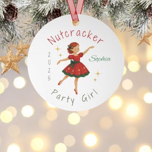 Peut inclure: Une décoration de Noël blanche et ronde, ornée d'un ruban rouge, représentant une ballerine en robe rouge avec des détails verts. L'ornement porte l'inscription "Nutcracker Party Girl Sophia 2025" en rouge et vert, avec des étoiles dorées.
