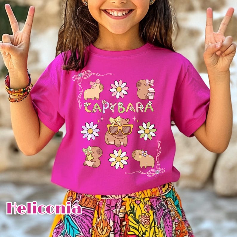 May include: A magenta t-shirt featuring a whimsical design with the word "CAPYBARA" and illustrations of capybaras and daisies. The shirt also has the word "Heliconia" at the bottom.