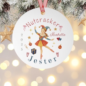 May include: A white Christmas ornament with a red ribbon features a ballerina in a colorful jester costume. The ornament has the words "Nutcracker" and "Jester", the name "Isabella", and the year "2025". Decorative stars and hearts surround the ballerina.