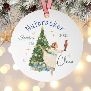 May include: A white round Christmas ornament with a red ribbon. The ornament features a ballerina holding a nutcracker, a Christmas tree, and the text "Nutcracker 2025". The ballerina is labeled "Clara" and the name "Sophia" is also on the ornament.