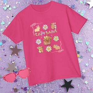 May include: A bright pink t-shirt featuring a whimsical design of capybaras, daisies, and the word "CAPYBARA". The capybaras are depicted with sunglasses, bows, and holding drinks. The shirt is surrounded by star-shaped confetti and heart-shaped sunglasses.