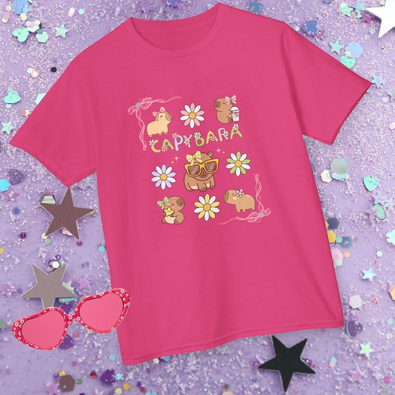 May include: A bright pink t-shirt featuring a whimsical design of capybaras, daisies, and the word "CAPYBARA". The capybaras are depicted with sunglasses, bows, and holding drinks. The shirt is surrounded by star-shaped confetti and heart-shaped sunglasses.