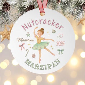以下が含まれることがあります： バレリーナのイラストが描かれた白い丸いクリスマスオーナメント。「Nutcracker」と「Marzipan」の文字が遊び心のあるフォントで書かれ、「Madeline」という名前と「2025」の年号が記されています。オーナメントは、お祝いのグラフィックで装飾されています。