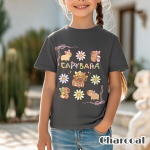May include: Charcoal gray t-shirt featuring a colorful capybara graphic. The design includes the word "CAPYBARA" and several capybara illustrations with flowers and accessories. The shirt is worn by a child.
