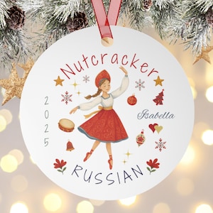 Könnte beinhalten: Runder, weißer Weihnachtsschmuck mit rotem Band. Der Schmuck zeigt eine Ballerina in russischer Tracht, den Text "Nutcracker Russian" und den Namen "Isabella". Dekorative Elemente sind Sterne, Schneeflocken und ein Weihnachtsbaum.