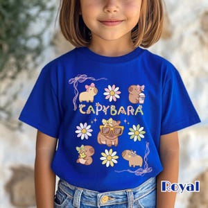 May include: Royal blue t-shirt featuring a colorful "CAPYBARA" design with cartoon capybaras and flowers. The capybaras are wearing bows and sunglasses. The shirt is made of a soft material.