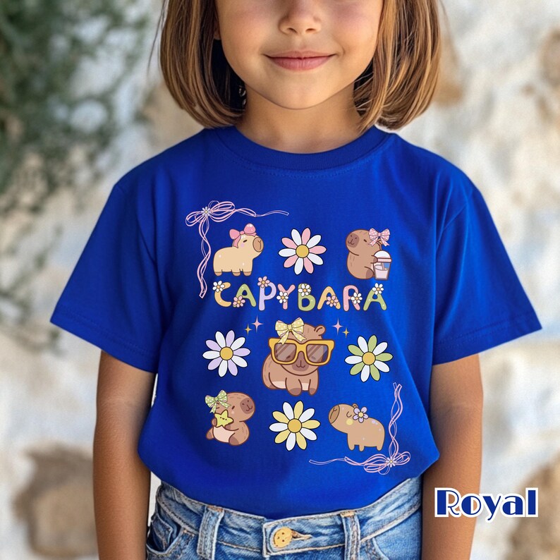 May include: Royal blue t-shirt featuring a colorful "CAPYBARA" design with cartoon capybaras and flowers. The capybaras are wearing bows and sunglasses. The shirt is made of a soft material.