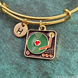 May include: Gold bangle bracelet with a charm of a vintage record player with a red heart on the record. The charm also has a small initial 'H' charm attached.