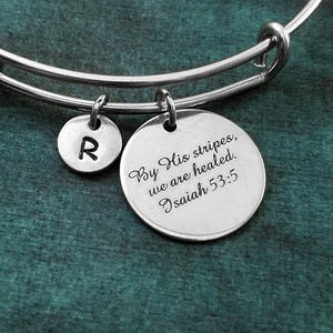 May include: A silver bangle bracelet with two charms. One charm is a round disc with the text "By His stripes, we are healed. Isaiah 53:5". The other charm is a round disc with the letter "R" engraved on it.