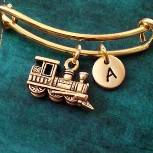 May include: Gold charm bracelet with a train charm and a letter A charm. The bracelet is on a teal background.