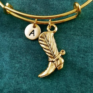 May include: Gold-colored bangle bracelet with a charm featuring a cowboy boot and a circular charm with the letter "A". The bracelet is displayed on a teal green surface. The text "MetalSpeakBangles" is visible at the bottom.
