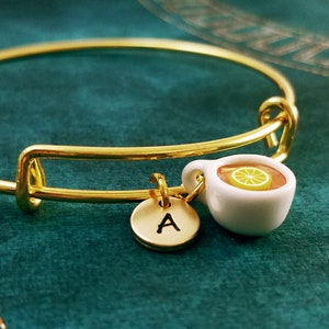 May include: A gold bangle bracelet with a charm of a white teacup with a lemon slice and a gold charm with the letter A.