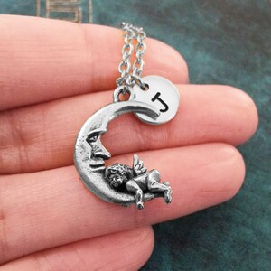 May include: A silver necklace with a crescent moon charm featuring a sleeping cherub and a round charm with the letter 'J'.