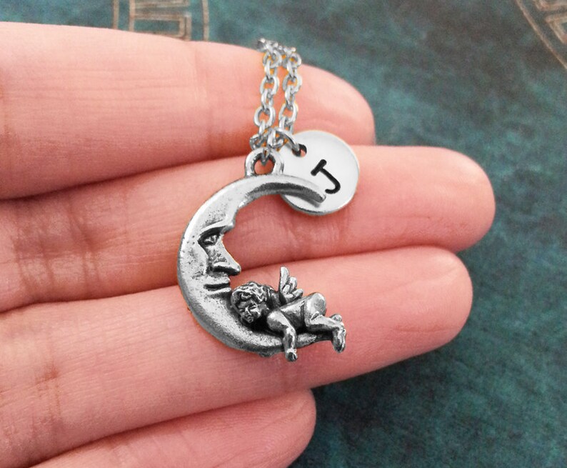 May include: A silver necklace with a crescent moon charm featuring a sleeping cherub and a round charm with the letter 'J'.