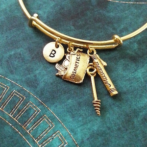 May include: Gold-colored expandable bracelet with cosmetic-themed charms. Charms include a round initial "B", a bag with the word "Cosmetics", a mascara tube, and a mascara wand. The bracelet is on a teal background.