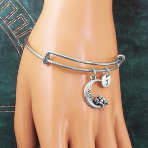 May include: Silver expandable bangle bracelet with a charm of a sleeping angel on a crescent moon and a silver disc charm with the letter "J" engraved on it.