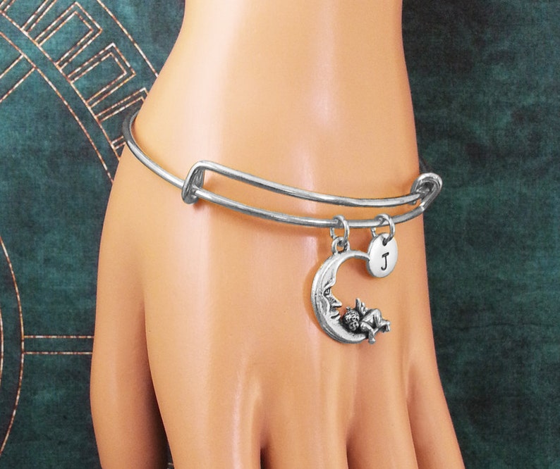 May include: Silver expandable bangle bracelet with a charm of a sleeping angel on a crescent moon and a silver disc charm with the letter "J" engraved on it.