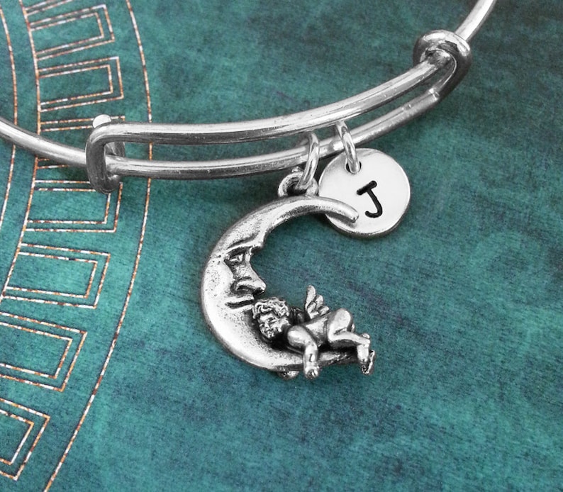 May include: Silver charm bracelet with a crescent moon charm featuring a sleeping cherub and a silver charm with the letter J.