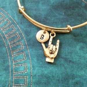 Könnte beinhalten: Goldenes Armkettchen mit einem Anhänger, auf dem "B" steht, und einem Anhänger in Form einer Hand, die das Zeichen für "Ich liebe dich" in Deutscher Gebärdensprache macht.