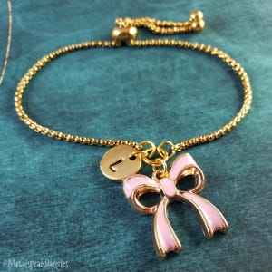 May include: Gold-toned bracelet with a pink bow charm and a small gold disc with the letter "L". The bracelet has a delicate chain design and a sliding clasp. The bow charm has a gold outline and pink enamel filling.