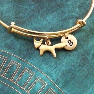 May include: Gold bangle bracelet with a fox charm and a round charm with the letter "B" engraved on it.