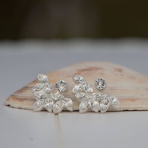 Pode incluir: Um par de brincos de prata com um design de folhas e cristais transparentes. Os brincos estão sobre uma superfície branca.