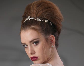 May include: A woman with a bouffant hairstyle wears a white, lace-like dress and a silver headband with small, clear stones. She has dark red lipstick and a serious expression.
