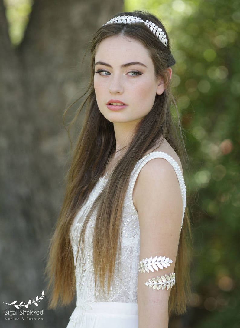 May include: A woman wearing a white lace dress with a silver leaf headband and matching silver leaf arm cuff.