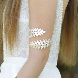 May include: A silver leaf-shaped arm cuff with a delicate design. The cuff is worn on a woman's arm, and the background is a white lace dress.