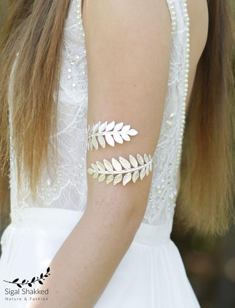 May include: A silver leaf-shaped arm cuff with a delicate design. The cuff is worn on a woman's arm, and the background is a white lace dress.