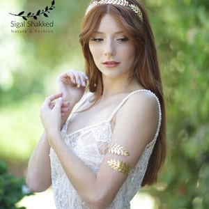 May include: A woman with long red hair wearing a white lace dress with a gold leaf headband and matching gold leaf arm cuff.