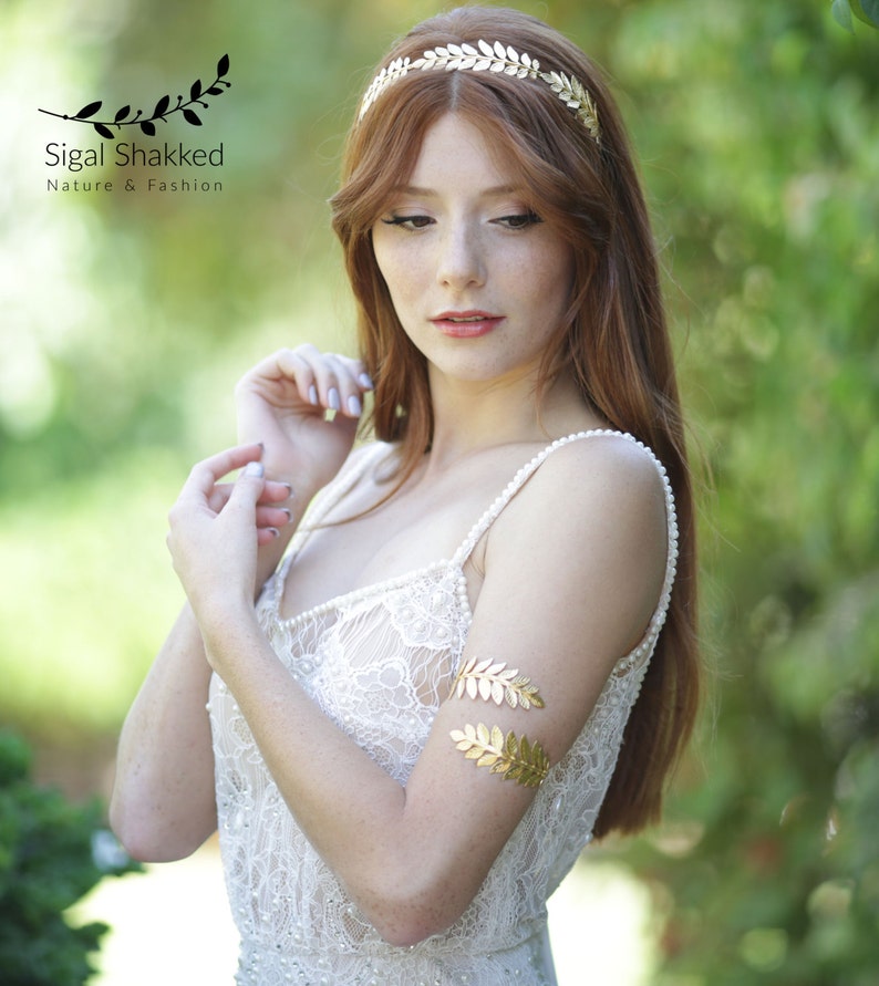 May include: A woman with long red hair wearing a white lace dress with a gold leaf headband and matching gold leaf arm cuff.