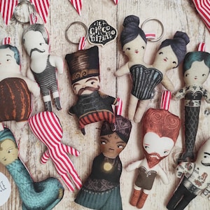 May include: A collection of 10 handmade fabric doll keyrings featuring various designs, including a strongman, a mermaid, a two-headed lady, and a bearded man. The dolls are all dressed in colourful outfits and have unique facial features. The keyrings are labelled "Il Circo Bizzarro" and "Le Bambolece".