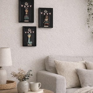 May include: Three framed wall art pieces featuring whimsical figures. Each framed piece has a black frame and a black background. The figures are colorful and detailed, with a vintage aesthetic. The art is displayed on a textured white wall.