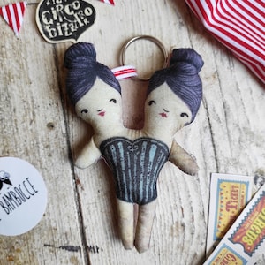 May include: A two-headed doll keychain with black hair, wearing a black and white striped swimsuit. The doll is made of fabric and has a whimsical, circus-themed design. The keychain is attached to a silver ring.