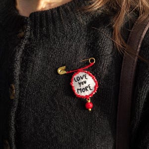 Op de afbeelding: Een goudkleurige veiligheidsspeld-broche met een ronde witte stoffen patch met de zwart geborduurde tekst LOVE you MORE. De patch is omrand met rood garen en heeft een kleine, bungelende rode kraal aan de onderkant, bevestigd op een zwarte trui.