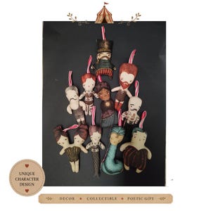 Op de afbeelding: Een verzameling van tien kleine stoffen ornamenten met geïllustreerde circusfiguren, waaronder een circusdirecteur, een zeemeermin en een figuur met twee hoofden. Elke zachte pop heeft een rood-wit gestreept lusje om op te hangen.
