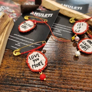 May include: Four handmade fabric pins with positive messages. The pins are red and white with gold safety pins. The messages are "Positive Vibes", "Let It Go", "Good Vibes", and "Love You More".