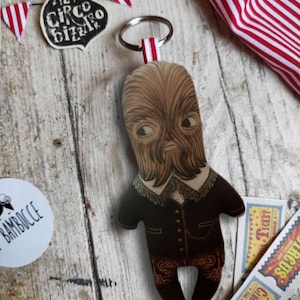 May include: A brown and black fabric keychain with a cartoon character wearing a black suit and brown pants. The character has a long beard and a mustache. The keychain is attached to a silver key ring. The background includes a wooden surface, red and white striped fabric, and a black and white sticker with the text "Il Circo Bizzarro".