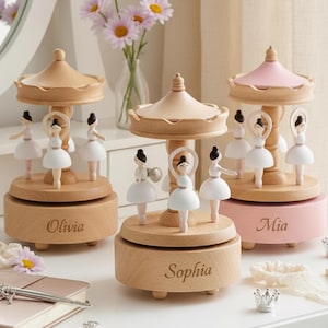 Może przedstawiać: Trzy drewniane pozytywki w kształcie karuzeli z figurkami baletnic. Jedna ma różowy wierzch, pozostałe są z naturalnego drewna. Każda podstawa jest grawerowana z imieniem: Olivia, Sophia i Mia. Figurki noszą białe tutu.