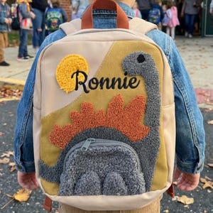 May include: A beige and tan backpack with a dinosaur design. The backpack features a yellow sun, a grey dinosaur, and the name "Ronnie" in black. It has a small front pocket and brown straps.
