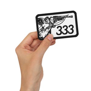 Könnte beinhalten: Ein rechteckiger schwarz-weißer Aufnäher mit einem Engel, der eine Trompete spielt, und der Zahl "333" in fetter schwarzer Schrift. Der Aufnäher wird in einer Hand vor weißem Hintergrund gehalten. Die Engel-Illustration ist detailliert und der Aufnäher hat einen schwarzen Rand.